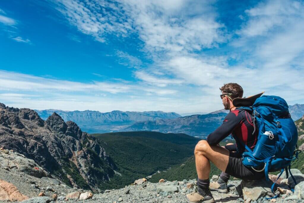 A man with a heavy backpack sits on a rock overlooking a mountain range